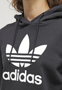 Svart hoodie med stor vit Adidas-logotyp med tre ränder och en treklöverdesign. Dragsnören vid halsen. Mjuk tygstruktur.