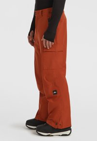 Orange waterproof snow pants feature a relaxed fit, multiple cargo pockets, adjustable waistband, and reinforced cuffs. Black hardware accents included.