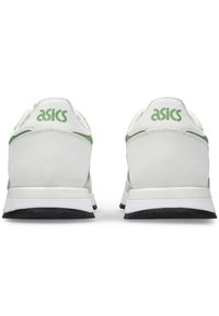 Par belih Asics superg z zelenimi detajli, gledano s hrbta, prikazuje logotip blagovne znamke na zadnjem delu in črne podplati.