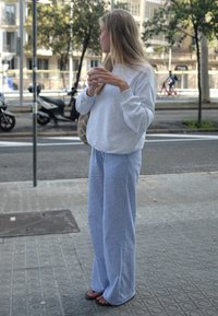 Femme aux longs cheveux blonds, portant un pantalon rayé bleu et un sweat-shirt gris, tenant une tasse tout en se tenant sur un trottoir en ville.