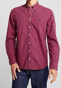 Camicia a righe rossa e blu con bottoni, maniche lunghe, chiusure a bottone bianche e un piccolo logo sul lato sinistro del petto. Tessuto di cotone.