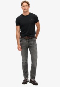 Camiseta negra ajustada, vaqueros grises con un leve desgaste, cinturón de cuero marrón y zapatos oscuros. Diseño sencillo sin patrones ni logotipos visibles.