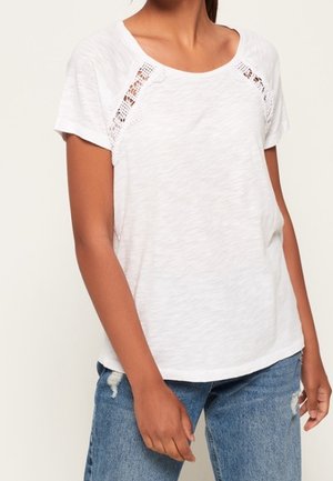 Mujer con una camiseta blanca de manga corta con detalle de encaje cerca de los hombros y vaqueros azules desgastados, de pie con los brazos relajados.