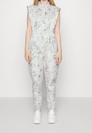 Bloemenpatroon jumpsuit in lichtblauwe stof, mouwloos ontwerp, met een kraag en elastische taille. Gecombineerd met witte sneakers.