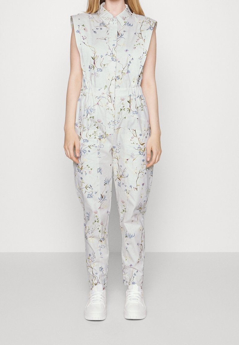 Floral-patterned jumpsuit in light blue fabric, sleeveless design, with a collar and elastic waist. Paired with white sneakers.