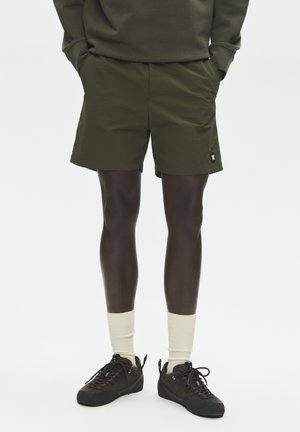 Persona con pantalones cortos verde oliva, calcetas blancas, zapatillas marrón oscuro y una camiseta verde oliva a juego, con las manos en los bolsillos.