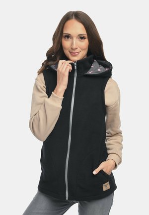 Frau trägt eine schwarze Fleeceweste mit Reißverschluss und sternbedrucktem Kapuzenfutter, beige lange Ärmel, graue Hose, lächelt und hält den Reißverschluss fest.