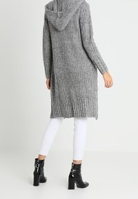 Szary długi kardigan z kapturem, wykonany z prążkowanego materiału, z otwartym przodem i rozcięciami po bokach, zestawiony z białymi obcisłymi spodniami i czarnymi botkami za kostkę.