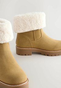 Bruna mocka ankelskor med krämfärgade fluffiga manschetter, tjocka gummisulor och kontrasterande sömmar längs kanten. Strukturerade och hållbara.