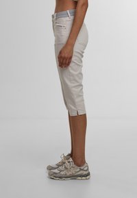 Beige Capri-Hosen mit einer taillierten Passform, ausgestattet mit einem grauen Bund, mittlerem Schnitt und seitlichen Schlitzen am Saum. Kombiniert mit hellen Sneakers.