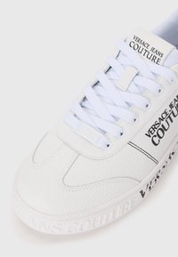 Baltos odinės sportbačiai su tekstūruota paviršiumi, apvaliu pirštu, plokščiomis baltomis raištelėmis ir juodu užrašu "VERSACE JEANS COUTURE" šone ir ant kulno.