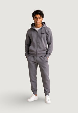 Ung mand stående med hænderne i lommerne iført grå lynlåshættetrøje, matchende joggingbukser, hvide sneakers og hvid t-shirt på lys baggrund.