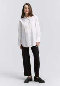 Camicia bianca a maniche lunghe con bottoni frontali e orlo arrotondato, abbinata a pantaloni larghi neri e scarpe con piattaforma nere.