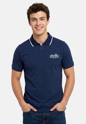 Jeune homme souriant portant un polo bleu marine à manches courtes avec un liseré blanc et un logo sur la poitrine, les mains dans les poches.