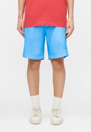 Osoba w jasnoniebieskich szortach, białych skarpetkach, kremowych tenisówkach i czerwonej koszulce, stojąca na tle jednolitego jasnego tła.