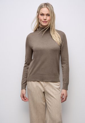 Blonde Frau trägt einen hellbraunen Rollkragenpullover und beigefarbene Hose, steht vor einer schlichten hellen Wand und schaut in die Kamera.