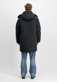Mann som står med ryggen til og har på seg en lang, svart hettejakke med dunfyll, falmede blå jeans og svarte sko mot en hvit bakgrunn.