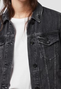 Černá denimová bunda se zapínáním na knoflíky, dvěma náprsními kapsami a límečkem. Nositá přes bílý tričko s texturovaným povrchem.