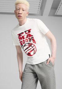 Hvid t-shirt med rød og sort grafisk tryk, der indeholder tekst og et basketballdesign. Grå bukser, casual fit og blød stofstruktur.