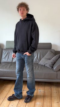 Must kapuutsiga pusa, lahtise lõikega, esiosas taskuga; kokku sobitatud siniste kulunud teksade ja tumepruunide kingadega. Seisab puidust põrandal.