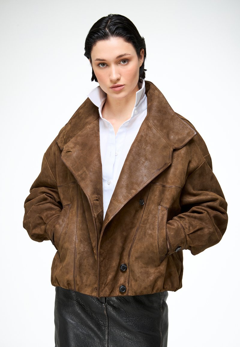 Fiatal nő rövid sötét hajjal, túlméretezett barna szarvasbőr dzsekit, fehér inget felhajtott gallérral és fekete bőr szoknyát visel, kezét zsebében tartva pózol.