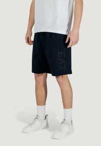 Pantaloni corti di cotone blu navy con elastico in vita, caratterizzati da una grafica logo grigia sulla gamba sinistra. Abbinati a sneakers bianche e calze alte.