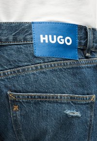 Μπλε τζιν παντελόνι με μικρή φθορά στην πίσω τσέπη και μια μπλε δερμάτινη ετικέτα ραμμένη πάνω της με την επιγραφή "HUGO".