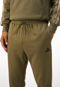 Olivza zaļas sporta bikses no mīksta auduma, ar elastīgu jostasvietu, auklu un melno Adidas logo uz augšstilba.