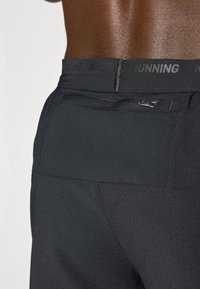 Schwarze Laufshorts mit einem strukturierten, perforierten Design, ausgestattet mit einer Reißverschlusstasche und einem elastischen Bund, der mit "NIKE RUNNING" geprägt ist.