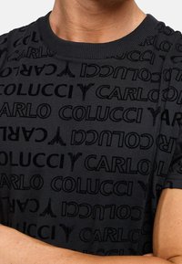 Černé bavlněné tričko s celoplošným vzorem názvu značky "CARLO COLUCCI" v různých orientacích a texturách. Design s kulatým výstřihem.
