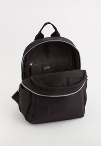 Mochila de nylon negra con forma redondeada, que cuenta con un asa superior, cremallera plateada y bolsillo interior. Textura suave con un diseño minimalista.