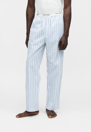 Hombre con pantalones sueltos de rayas verticales blancas y azules con cintura de la marca Calvin Klein y una camiseta blanca, descalzo sobre fondo liso.