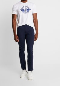 Bílý bavlněný tričko s modrou grafiku "Dockers", kombinované s námořnickými slim-fit kalhotami a bílými teniskami, předvádějící neformální outfit.