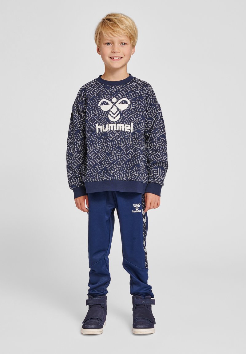 Blond dreng står smilende iført navyblå hummel sweatshirt med logo og matchende joggebukser samt mørkeblå sneakers mod en ensfarvet grå baggrund.