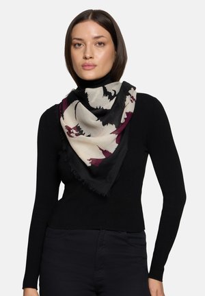 Femme portant un col roulé noir et un pantalon foncé avec une grande écharpe noire, blanche et bordeaux à motifs drapée autour de son cou.