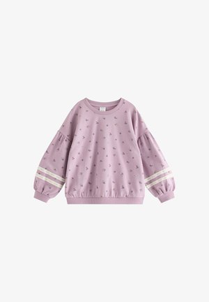 Jeune fille debout souriante, portant un sweat et un pantalon lilas à motifs floraux assortis avec des bandes en dentelle et un élastique à cheveux assorti.