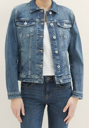 Personne portant une veste en jean bleu moyen par-dessus un haut blanc et un jean bleu foncé, les mains détendues le long du corps.