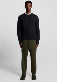 Schwarzer gerippter Strickpullover mit langen Ärmeln, kombiniert mit grünen gerade geschnittenen Hosen und schwarzen Schnürschuhen, stehend vor einem neutralen Hintergrund.