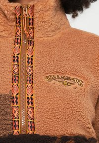 Barna polár pulóver magas gallérral, narancs és fekete mintás cipzárral, valamint a mellrészen hímzett logóval.