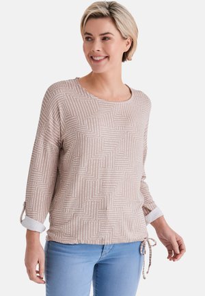 Frau lächelt, trägt ein beiges Langarmshirt mit weißem geometrischem Punktemuster und hellblaue Jeans, steht vor einem einfarbigen Hintergrund.