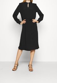 Schwarzes Kleid mit langen Ärmeln, Schlüssel-Ausschnitt und Taillenkrawatte. Hergestellt aus einem weichen Stoff, hat es einen fließenden Rock und eine dezente Textur.
