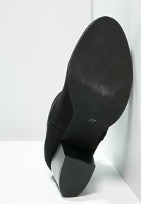 Czarny zamszowy but na wysokim obcasie z teksturowanym, zwężającym się obcasem. Podeszwa jest gładka, z widocznym oznaczeniem rozmiaru 37.