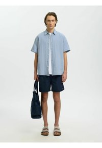 Jeune homme portant une chemise à manches courtes rayée bleue, un short bleu marine, des sandales beige, tenant un sac cabas bleu marine, debout devant un fond blanc uni.
