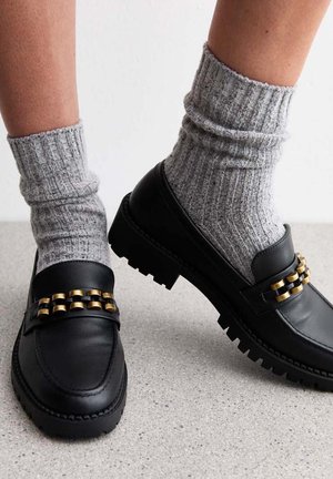 Black leather loafers with a gold chain accent, paired with grey ribbed socks. The shoes feature a chunky sole and rounded toe.