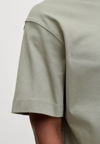 Olivgrünes Baumwoll-T-Shirt mit kurzen Ärmeln an einer Person mit dunkler Haut, zeigt detaillierte Nähte am Ärmel und an der Schulter.