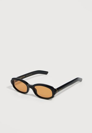 Črne ovalne sončna očala z jantarjevimi lečami in tankimi okvirji, položena na belo površino, z nežnim zlatim napisom na straneh.