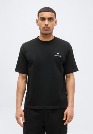 Jeune homme aux cheveux courts portant un t-shirt noir uni avec le logo « snow peak » sur la poitrine gauche, debout sur un fond blanc.
