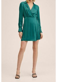 Robe en satin teal à manches longues, col à revers et fermeture par lien à la taille. Texture lisse et longueur courte. Sandales noires à brides.