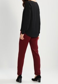 Blouse noire à manches longues avec un col arrondi, associée à un pantalon slim bordeaux et des chaussures à talons noires. Tissu lisse, coupe décontractée.