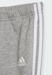 Pantaloni sportivi grigi con elastico in vita, caratterizzati da strisce verticali bianche e grigie scure ai lati e il logo Adidas.